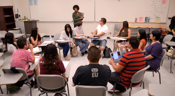 Students at desks looking at a teacher holding a book