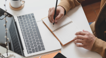 Person sitting at desk writing in spiral notebook in front of a laptop screen